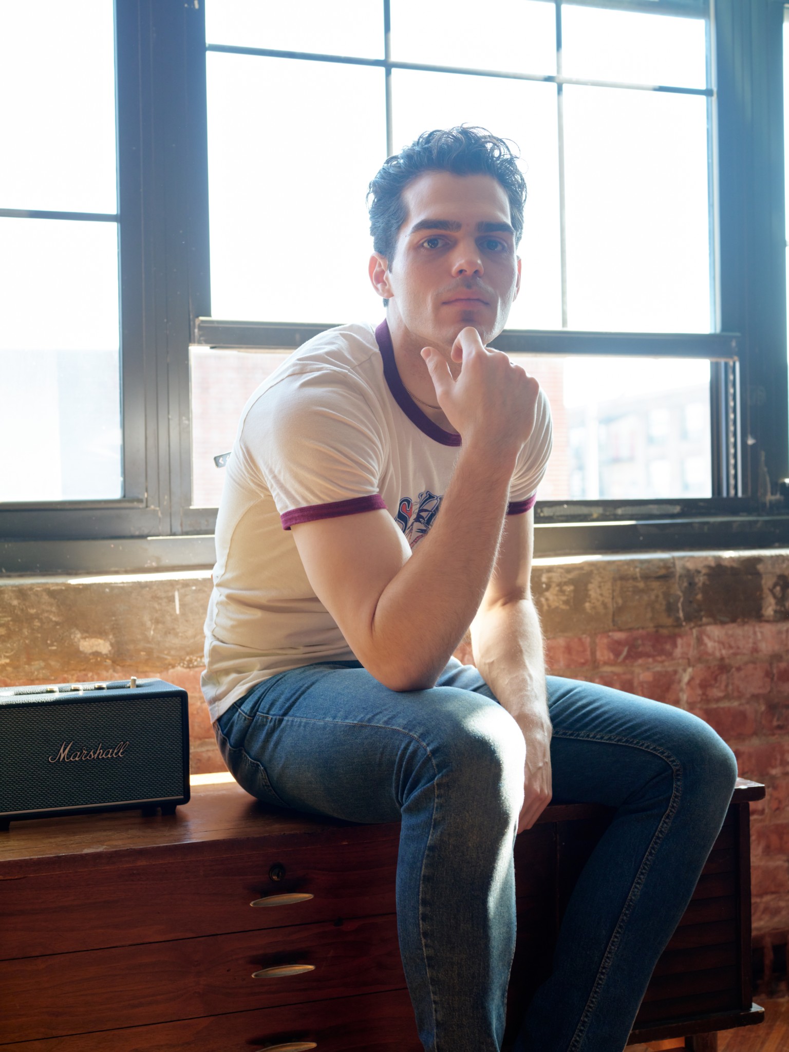 Man seated beside an amplifier in a bright, backlit studio window.